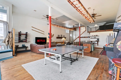 A spacious living area with a ping pong table, cozy seating, and a TV displaying Netflix, featuring wooden floors and bright decor.